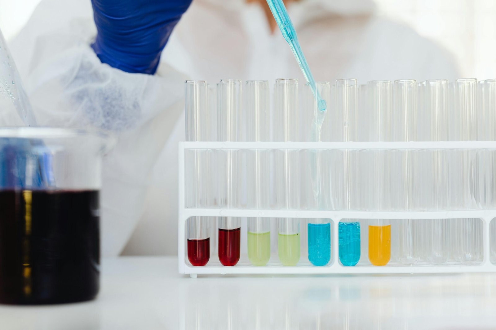 Scientist in protective gear using a dropper to fill test tubes with colorful liquids in a laboratory.